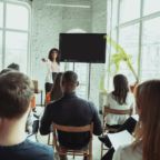 Female african-american speaker giving presentation in hall at workshop. Audience or conference hall. Rear view of participants in audience. Conference event, training. Education, diversity, inclusive concept.